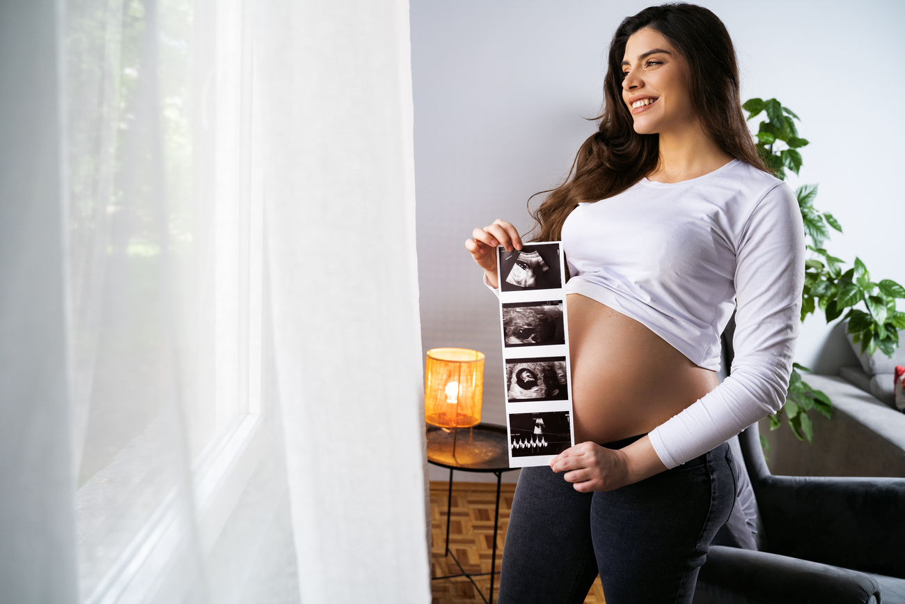 Amazing young pregnant woman standing next to window with ultrasound images smiling